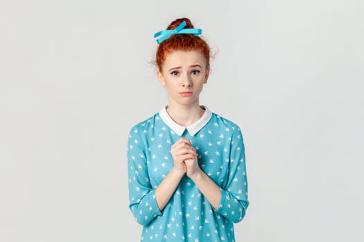 Portrait of hopeful beautiful ginger woman with bun hairstyle, standing with hands together, pleading, asking to forgive, wearing blue dress. Indoor studio shot isolated on gray background.