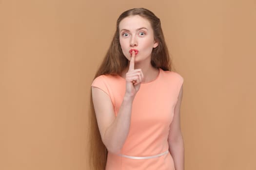 Portrait of attractive winsome woman with long hair showing shh gesture, keep silence, looking at camera with strict expression, wearing elegant dress. Indoor studio shot isolated on brown background.
