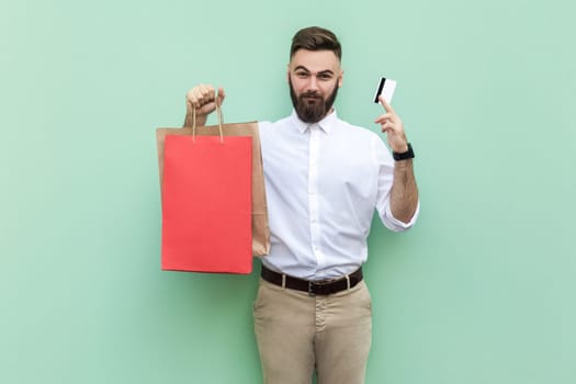 Portrait of smiling self-confident businessman wearing white shirt showing to camera red shopping bag and credit card for paying for purchases. Indoor studio shot isolated on light green background.