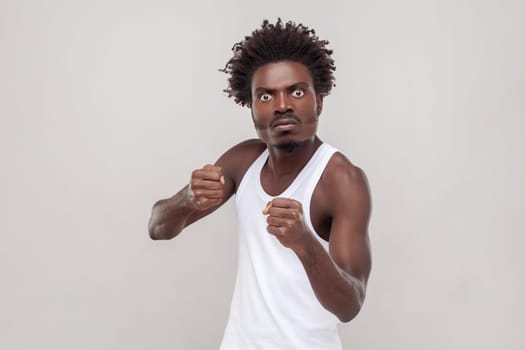 Displeased impassionate man fighting with someone, looking at camera, clenched fists, has furious facial expression. wearing white T-shirt. Indoor studio shot isolated on gray background.