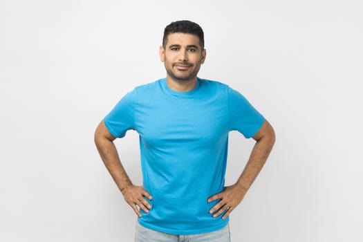 Portrait of happy delighted optimistic unshaven man wearing blue T- shirt standing looking at camera, expressing positive emotions, keeps hands on hips. Indoor studio shot isolated on gray background.