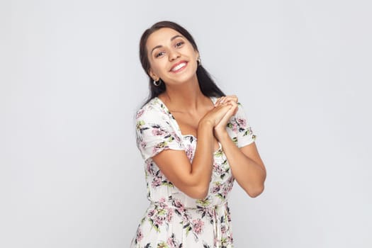 Portrait of cute charming beautiful woman wearing white dress looking at camera with toothy smile, keeps hands together, admires. Indoor studio shot isolated on gray background.