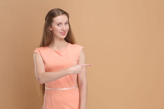Portrait of smiling positive friendly woman with long hair pointing aside at advertisement area, copy space for promotion, wearing elegant dress. Indoor studio shot isolated on brown background.