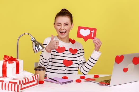 Smiling woman holding like counter template sitting at workplace covered heart sticks, making post in social network, showing thumb up. Indoor studio studio shot isolated on yellow background.