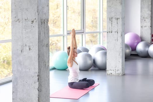 Side view portrait of sporty relaxed woman practicing yoga, doing Sukhasana exercise, Easy Seat pose raising arms up, lifting hands, working out in fitness club. Indoor shot with window on background.