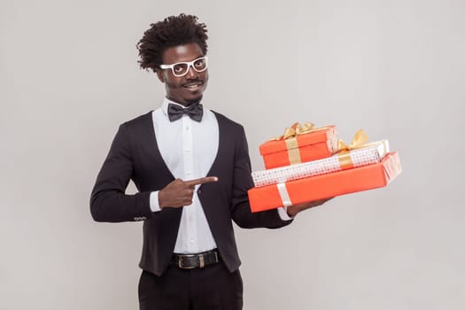 Portrait of attractive positive young adult man in glasses pointing at three present boxes in his hands, looking at camera, wearing shirt and tuxedo. Indoor studio shot isolated on gray background.