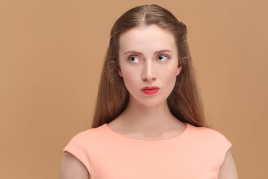 Portrait of attractive winsome woman with long hair and red lips, wearing elegant dress, looking away with serious confident expression. Indoor studio shot isolated on brown background.