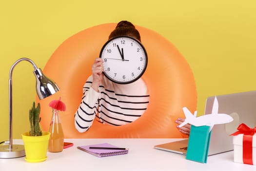 Portrait of unknown anonymous office employee in rubber ring on neck sitting at workplace with laptop, hiding face with wall clock. Indoor studio studio shot isolated on yellow background.