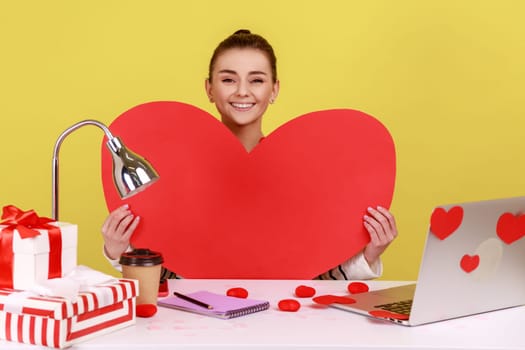 Happy woman holding big red paper heart and smiling, sitting at workplace with laptop in sticky hearts, showing her love. Indoor studio studio shot isolated on yellow background.