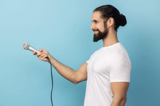 Side view of man with beard wearing white T-shirt holding microphone, making interview and asking opinion, discussing important topics. Indoor studio shot isolated on blue background.