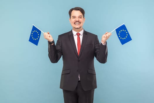 Portrait of positive man with mustache standing holding EU flags and looking at camera with toothy smile, wearing black suit with red tie. Indoor studio shot isolated on light blue background.