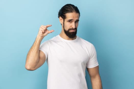 Man with beard wearing white T-shirt discusses something very small, shapes something very tiny, purses lips, asks tiny object, makes small gesture. Indoor studio shot isolated on blue background.