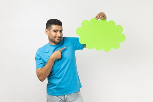 Cheerful unshaven man wearing blue T- shirt standing pointing at speech or think green bubble, empty communication cloud, looking at camera with smile. Indoor studio shot isolated on gray background.