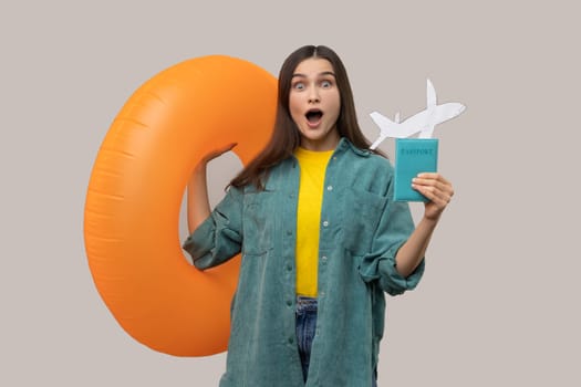 Portrait of amazed shocked woman holding orange rubber ring, passport document and paper airplane, traveling abroad, wearing casual style jacket. Indoor studio shot isolated on gray background.