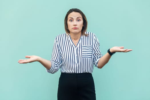 Uncertain clueless attractive middle aged woman wearing striped shirt doesn't know correct answer, looking at camera, shrugging shoulders. Indoor studio shot isolated on light blue background.