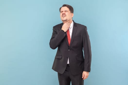 Man with mustache standing feels unwell, keeps hand on neck, has inflammation of throat, suffers from suffocation, wearing black suit with red tie. Indoor studio shot isolated on light blue background