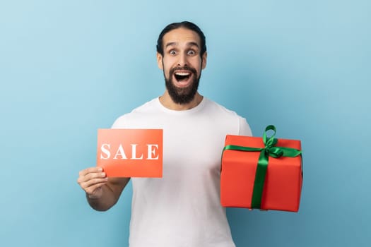 Portrait of amazed surprised man with beard wearing white T-shirt holding red gift box with green ribbon and showing sale coupon, huge sales. Indoor studio shot isolated on blue background.