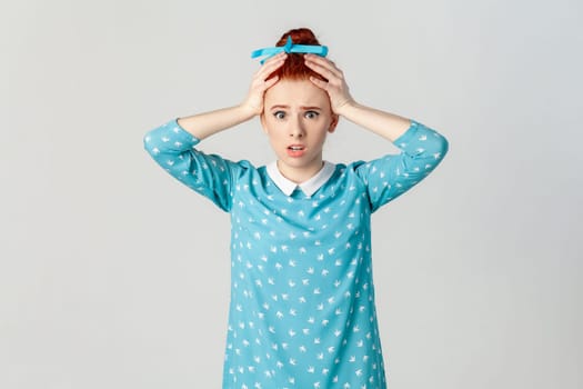 Portrait of astonished scared red haired woman with hair bun, standing with hands on her head, sees something shocked, wearing blue dress. Indoor studio shot isolated on gray background.