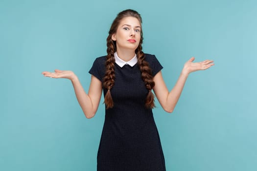 Puzzled uncertain young woman with braids spreads hands aside, shrugging shoulders, don't know how to make a decision, wearing black dress. woman Indoor studio shot isolated on blue background.