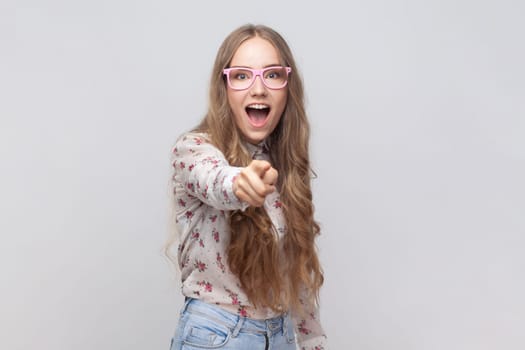 Portrait of surprised woman in glasses with wavy blond hair looking with big eyes and open mouth, pointing finger at camera, suspecting you. Indoor studio shot isolated on gray background.