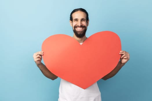 Portrait of delighted man with beard wearing white T-shirt holding big red heart in hands, looking at camera with positive, expressing love. Indoor studio shot isolated on blue background.