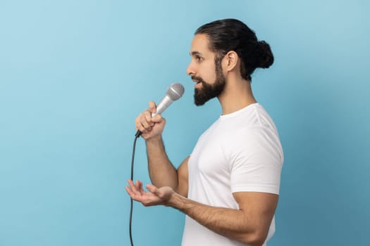 Side view of serious man with beard wearing white T-shirt, journalist holding microphone, discussing problems, reporting. Indoor studio shot isolated on blue background.