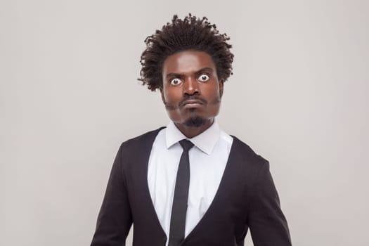 Portrait of man with Afro hairstyle looks with angry strict expression gazes directly at camera, has negative emotions, wearing white shirt and tuxedo. Indoor studio shot isolated on gray background.