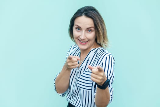 Portrait of satisfied attractive joyful middle aged woman wearing striped shirt pointing to camera with fingers, choosing you, making choice. Indoor studio shot isolated on light blue background.