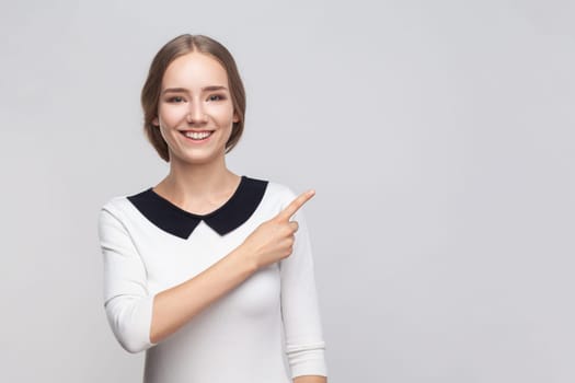 Portrait of pretty smiling optimistic woman wearing white dress pointing finger aside at advertisement area, copy space for promotion. Indoor studio shot isolated on gray background.