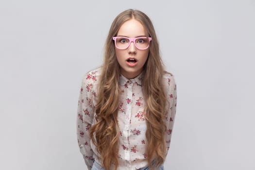 Portrait of astonished surprised woman in glasses with long blond hair looking with shocked expression at camera, keeping mouth widely open. Indoor studio shot isolated on gray background.