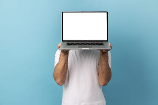 Portrait of man with beard wearing white T-shirt hiding his face behind laptop with blank display for advertisement or promotional text. Indoor studio shot isolated on blue background.