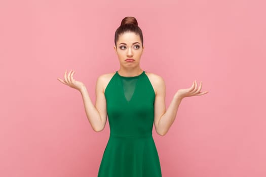Portrait of confused unsure woman with bun hairstyle standing with spread hands, looking at camera with uncertain expression, wearing green dress. Indoor studio shot isolated on pink background.