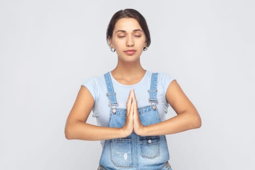 Portrait of calm relaxed young adult woman wearing denim overalls standing palm hands and doing yoga meditation with closed eyes. Indoor studio shot isolated on gray background.