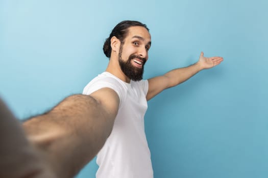 Portrait of king friendly man with beard wearing white T-shirt looking at camera with smile, making welcome gesture, taking selfie POV. Indoor studio shot isolated on blue background.