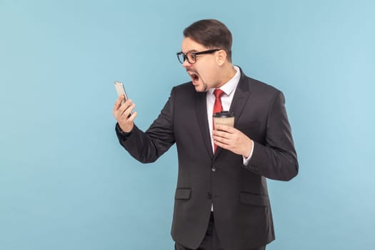 Portrait of scared shocked man standing using cell phone, looking at screen, screaming, holding cup of coffee, wearing black suit with red tie. Indoor studio shot isolated on light blue background.