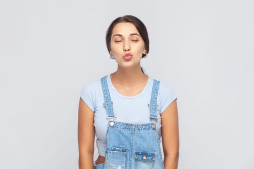 Portrait of attractive beautiful young adult woman wearing denim overalls standing with closed eyes, expressing love and sending air kiss. Indoor studio shot isolated on gray background.