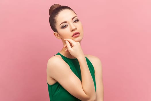 Portrait of feminine attractive beautiful woman with bun hairstyle, keeps hand under chin, looking at camera with passion, wearing green dress. Indoor studio shot isolated on pink background.
