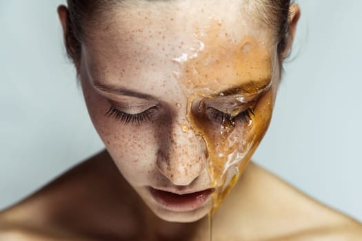 Closeup portrait of adorable winsome young brunette woman with freckles and honey mask, standing with closed eyes or looking down. Indoor studio shot isolated on gray background.