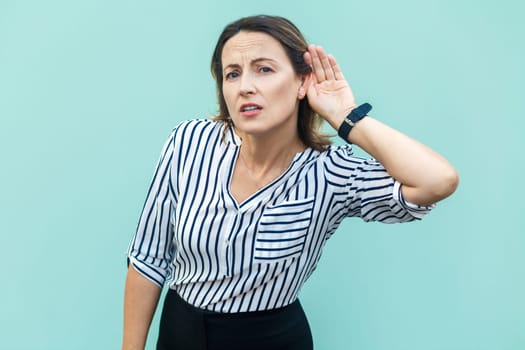 Portrait of curious beautiful middle aged woman wearing striped shirt standing with hand near ear, listening to gossips or confident information. Indoor studio shot isolated on light blue background.