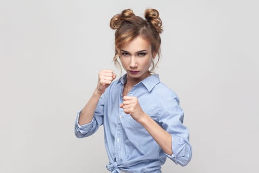 Woman having serious and confident look keeping fists clenched in front of her defencing herself from offence and bad treatment, wearing blue shirt. Indoor studio shot isolated on gray background.