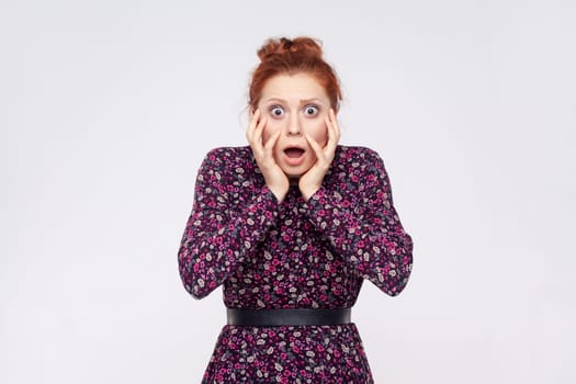 Portrait of shocked amazed astonished ginger woman wearing dress keeps jaw dropped and hands on cheeks, cannot believe in amazing news. Indoor studio shot isolated on gray background.