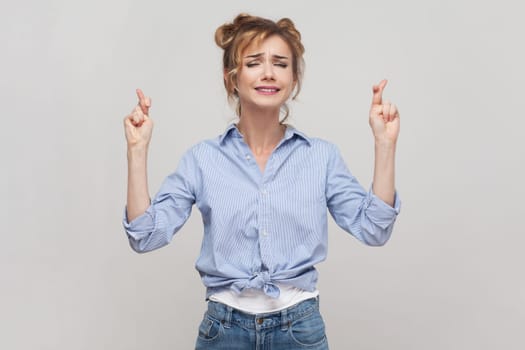 Cheerful blonde woman awaiting for miracle, praying and hopes dreams come true, crosses fingers for good luck, closes eyes, wearing blue shirt. Indoor studio shot isolated on gray background.