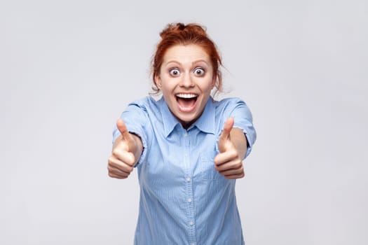 Portrait of positive redhead woman wearing blue shirt showing thumbs up, looking at camera with open mouth, surprised with excellent result. Indoor studio shot isolated on gray background.