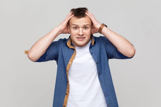 Portrait of scared attractive handsome teenager boy wearing blue shirt keeping hands on head and looking frighten with frightened eyes. Indoor studio shot isolated on gray background.