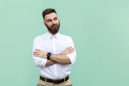 Portrait of young adult bearded businessman wearing white shirt standing with folded arms and looking away with smile and dreaming expression. Indoor studio shot isolated on light green background.