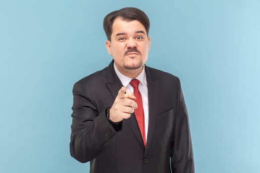 Portrait of sad upset man with mustache standing indicating at you, looking at camera with frowning face, wearing black suit with red tie. Indoor studio shot isolated on light blue background.