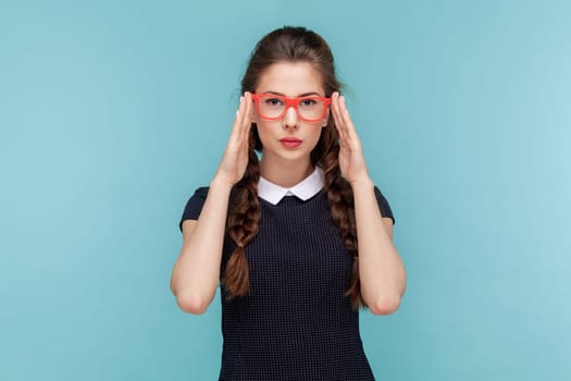 Portrait of serious attractive woman with braids stands keeps hands on frame of red glasses, looking at camera with attention, wearing black dress. woman Indoor studio shot isolated on blue background