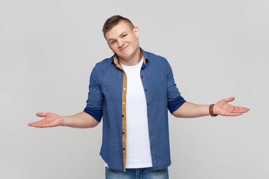 Portrait of open hearted generous teenager boy wearing blue shirt sharing opening hands looking at camera with kind smile, greeting and regaling. Indoor studio shot isolated on gray background.