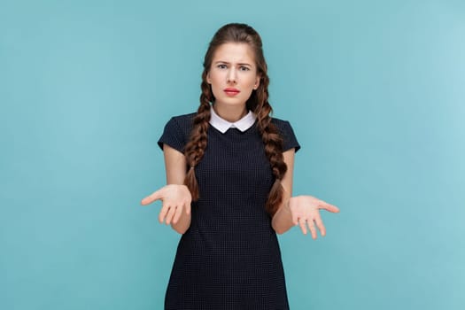How could you. Portrait of sad annoyed woman with braids standing with spread hands, being irritated, asking what, wearing black dress. woman Indoor studio shot isolated on blue background.