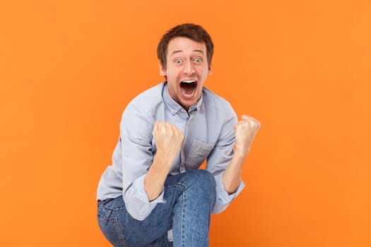 Portrait of excited amazed young adult man standing screaming happily, clenched fists, yelling with excitement, wearing light blue shirt. Indoor studio shot isolated on orange background.